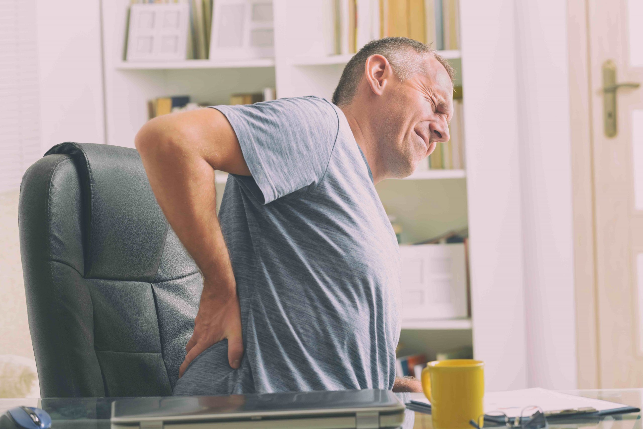 Man in home office suffering from low back pain sitting at the desk with notebook, papers and other objects